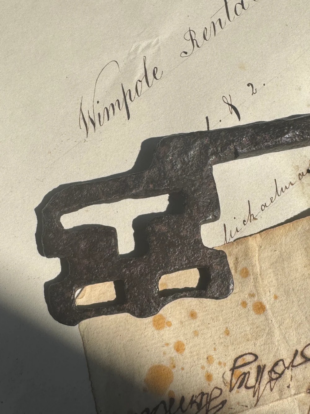 A hand-forged dark wrought iron key featuring a circular bow, a straight shaft, and a cut bit resting on manuscript paper. View IV