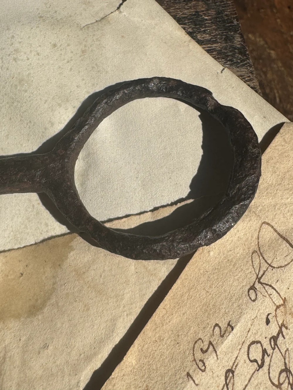 A hand-forged dark wrought iron key featuring a circular bow, a straight shaft, and a cut bit resting on manuscript paper. View V