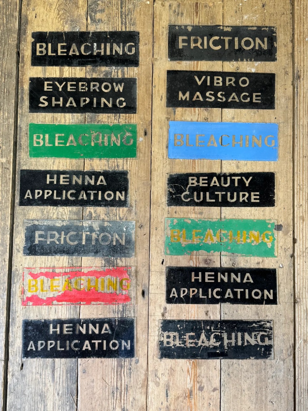 Fourteen rectangular reverse-painted glass signs detailing vintage beauty treatment terms in metallic lettering on painted backgrounds.