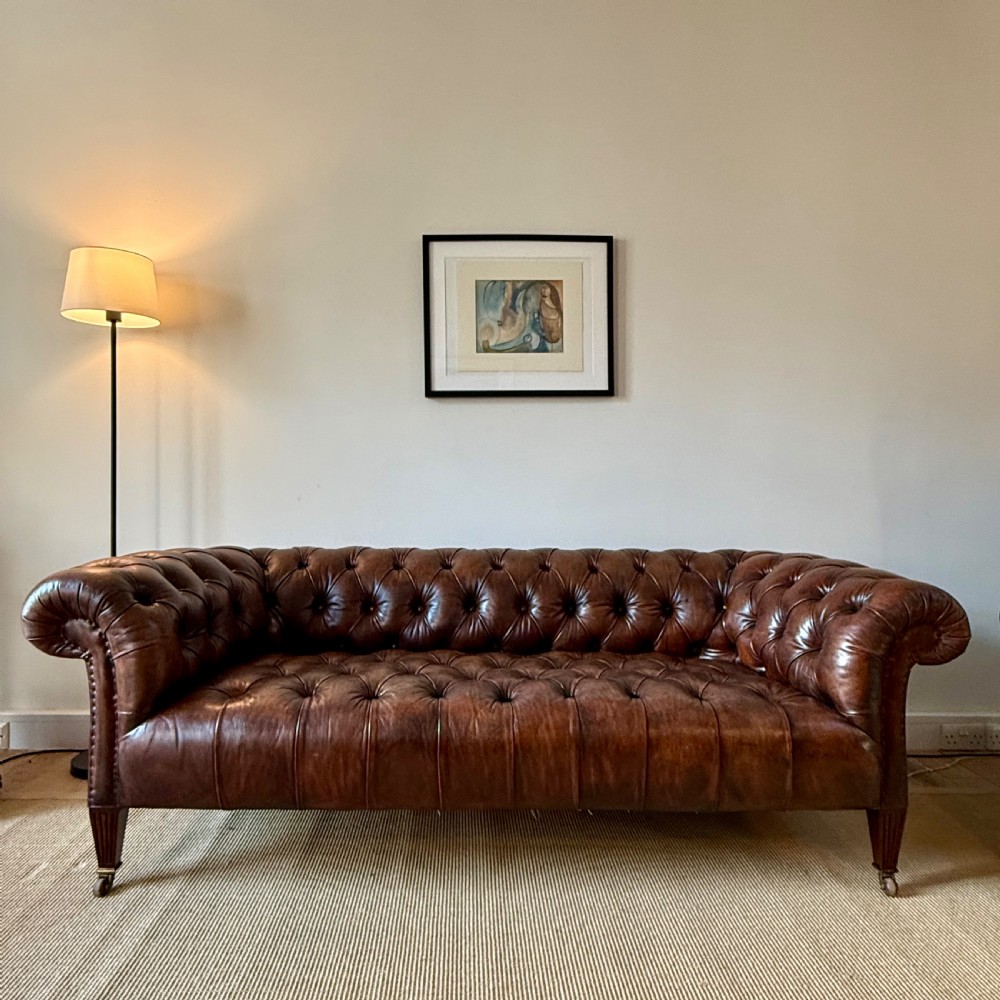 A 19th-century brown hand-dyed leather Chesterfield sofa featuring deep button tufting, rolled arms, and reeded wooden legs on brass castors. View II
