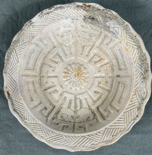 fig 1. A circular off-white antique ceramic dish featuring a scalloped rim, central meander pattern, and subtle marine mineral deposits.