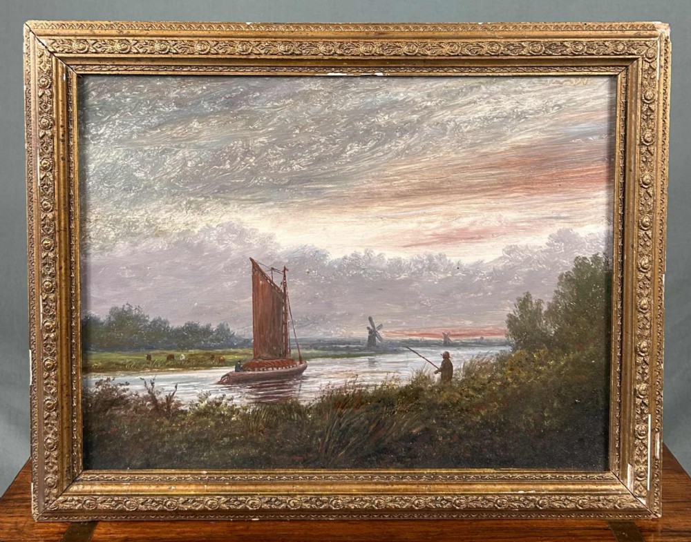 A framed oil painting featuring a river landscape in the Norfolk Broads with a sailboat, a fisherman on the bank, and a windmill under a textured cloudy sky.