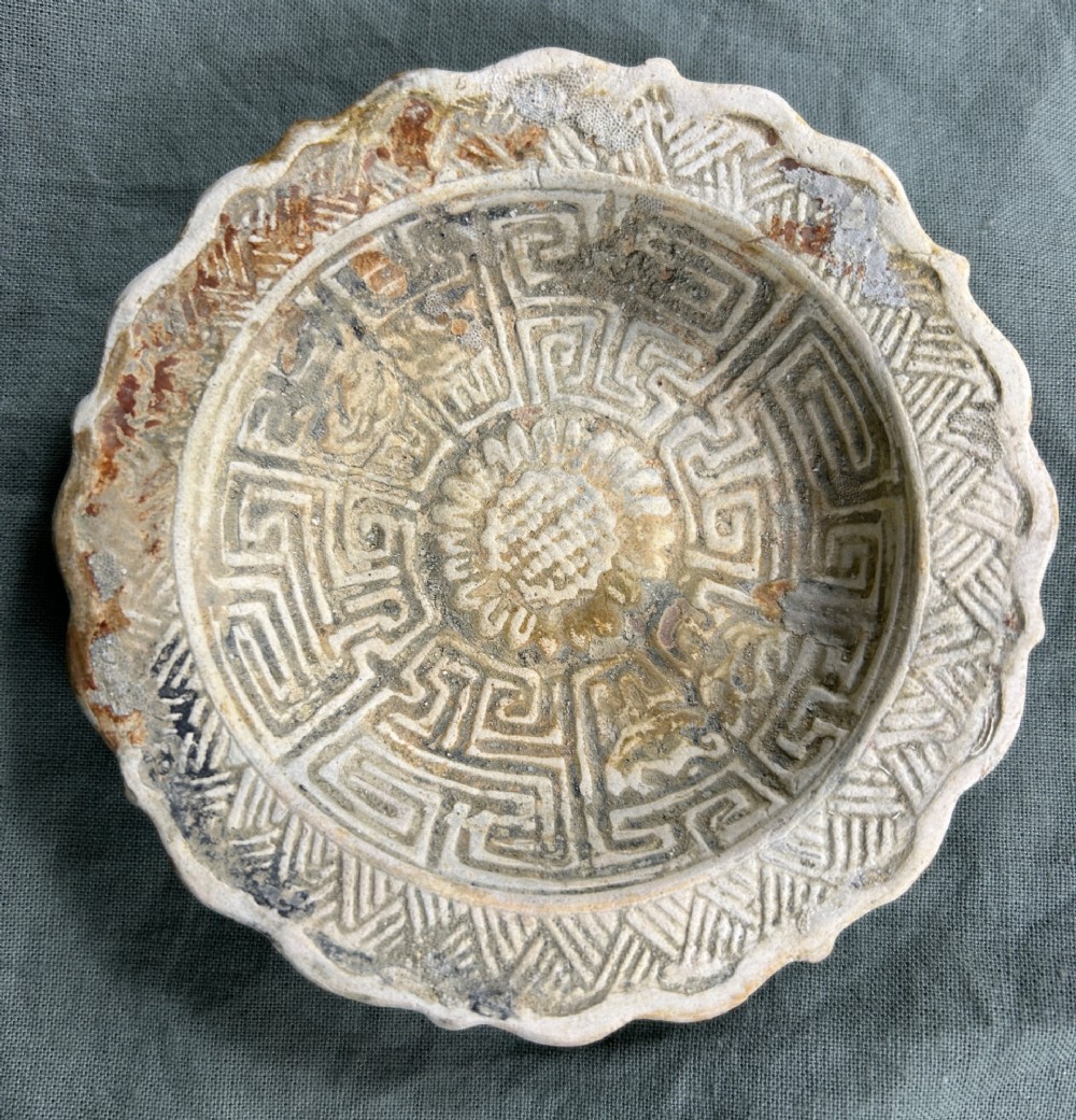 A circular off-white antique ceramic dish featuring a scalloped rim, central meander pattern, and subtle marine mineral deposits.
