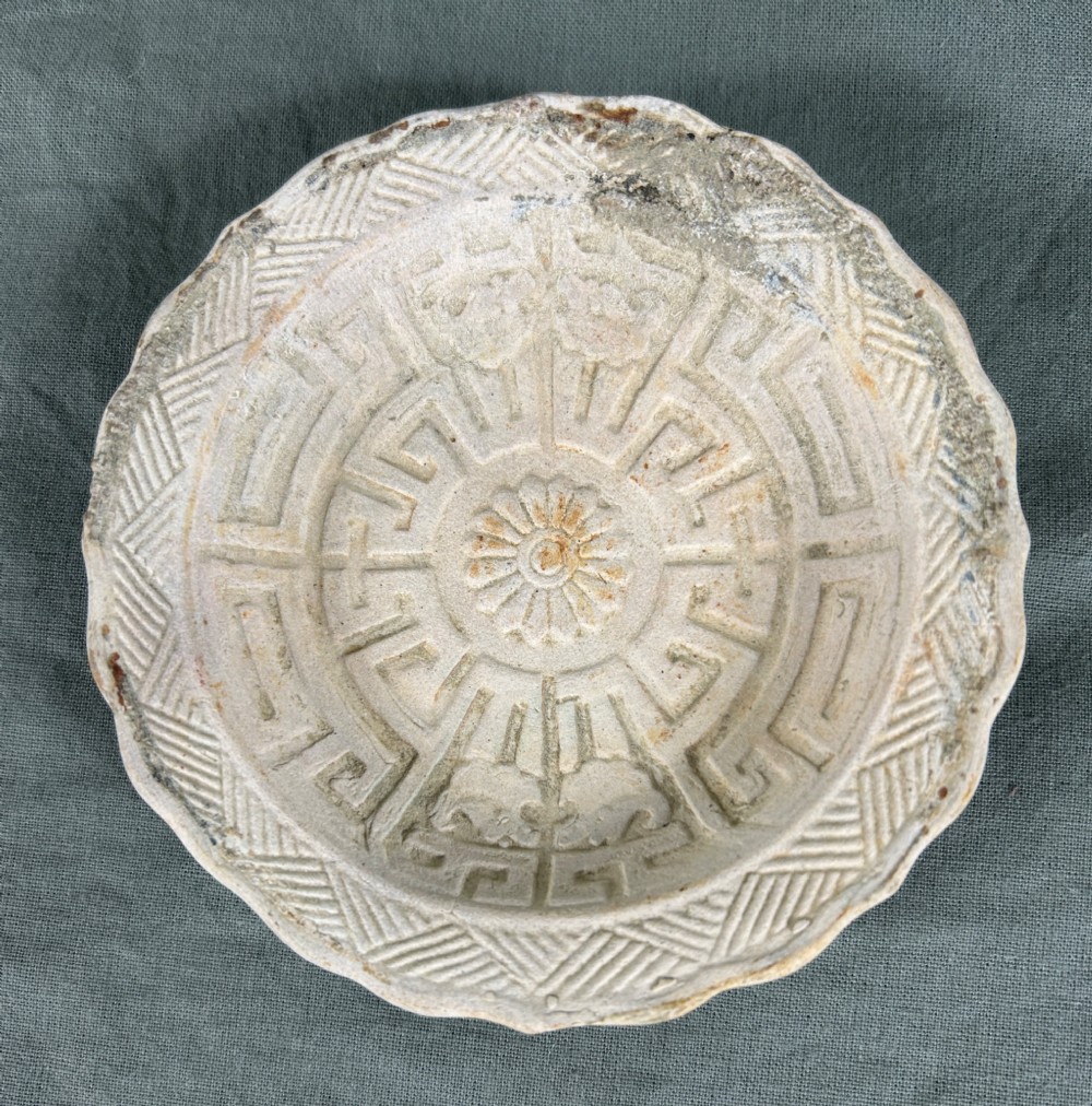 A circular off-white antique ceramic dish featuring a scalloped rim, central meander pattern, and subtle marine mineral deposits.