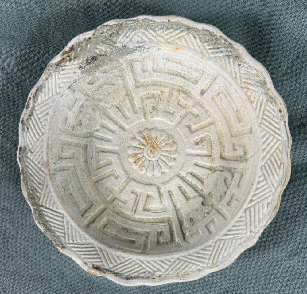 A circular off-white antique ceramic dish featuring a scalloped rim, central meander pattern, and subtle marine mineral deposits. View V