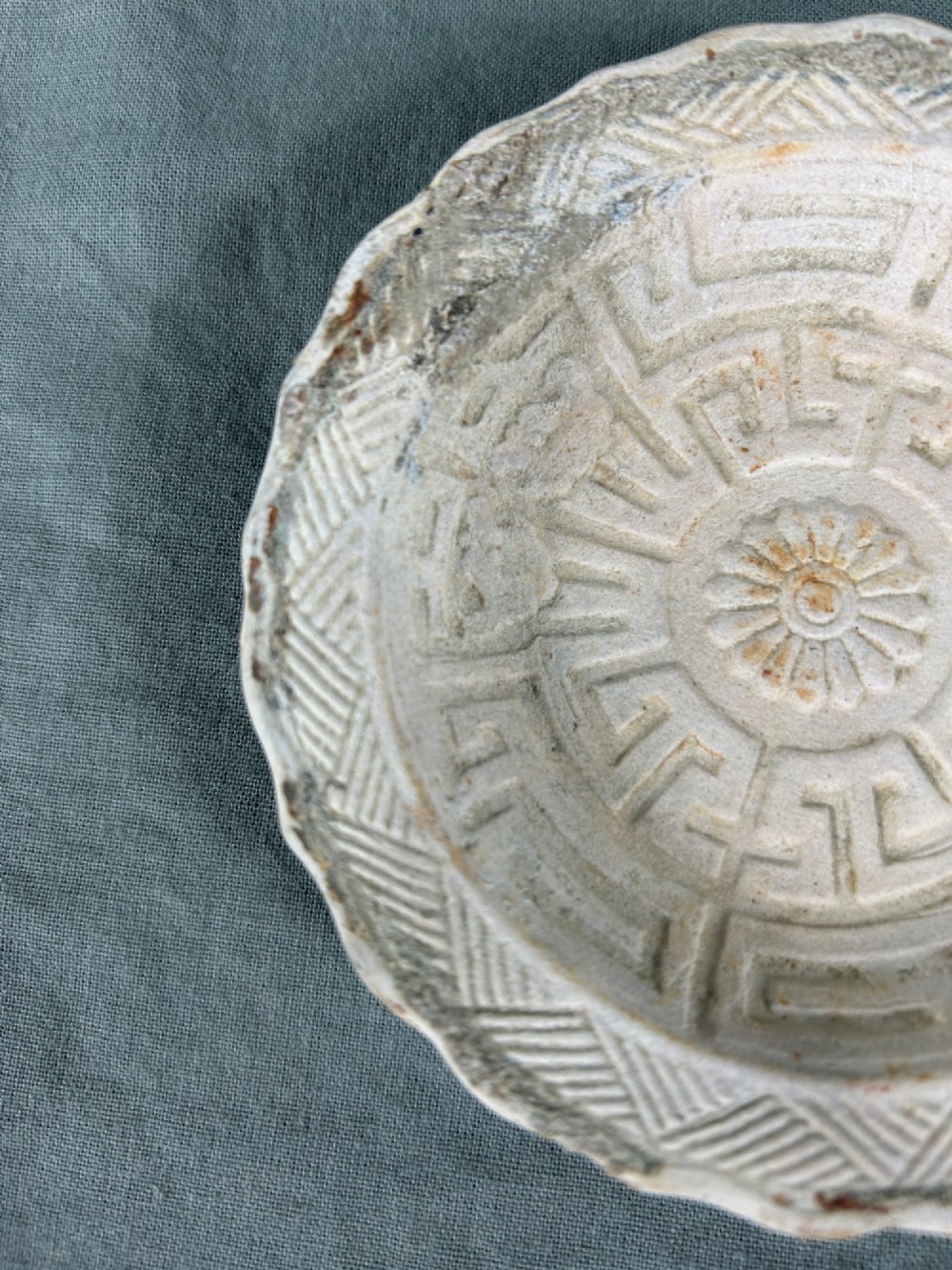 A circular off-white antique ceramic dish featuring a scalloped rim, central meander pattern, and subtle marine mineral deposits. View VI