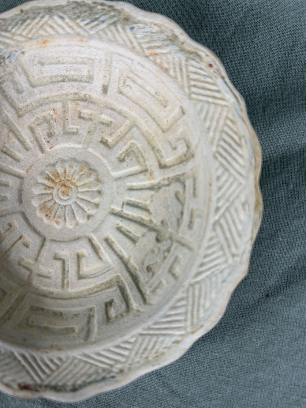 A circular off-white antique ceramic dish featuring a scalloped rim, central meander pattern, and subtle marine mineral deposits. View VII
