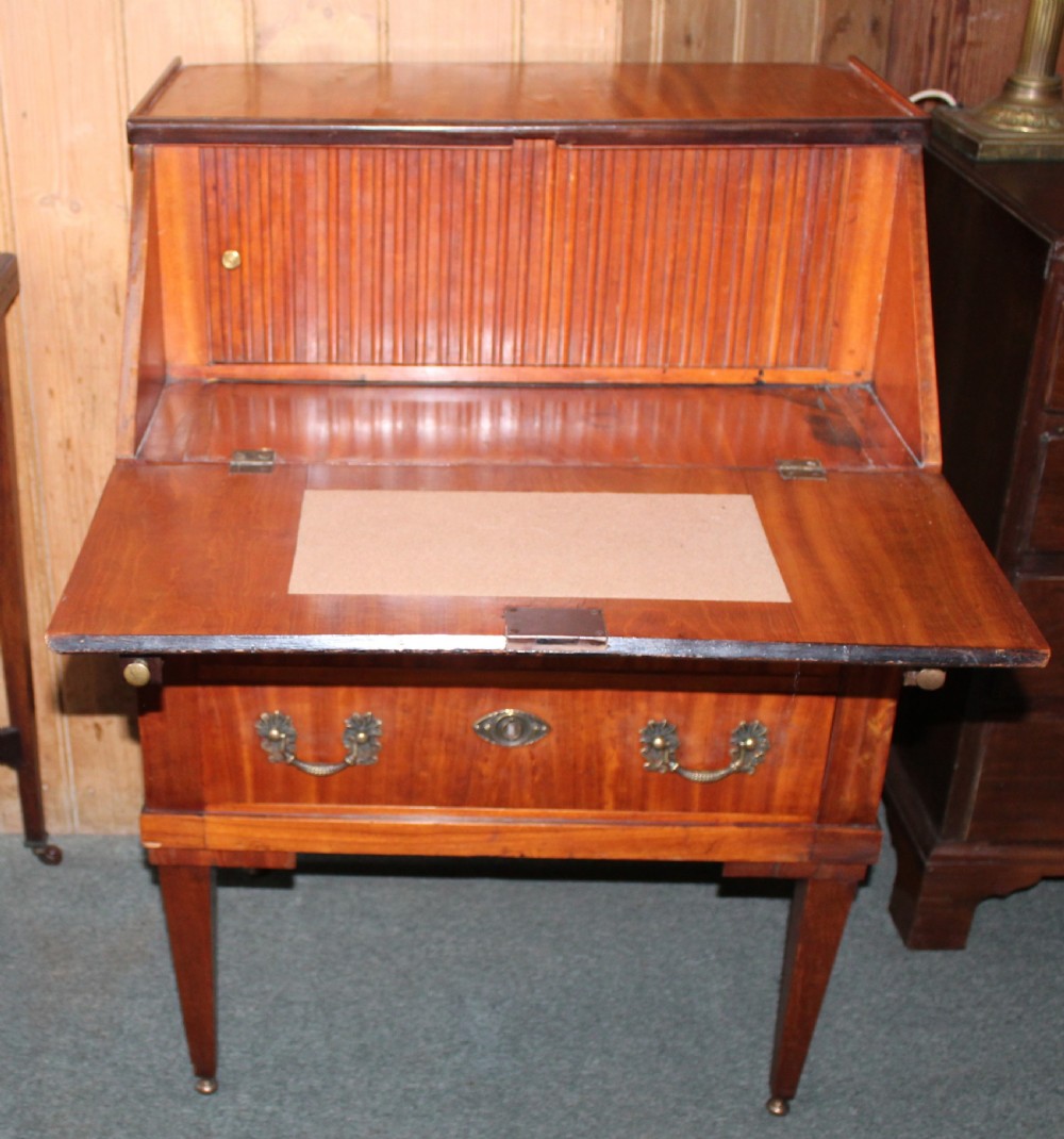 An Edwardian satinwood writing bureau featuring a sloping fall-front lid, two lower drawers with brass drop handles, dark crossbanded edging, and square tapered legs on castors. View VI