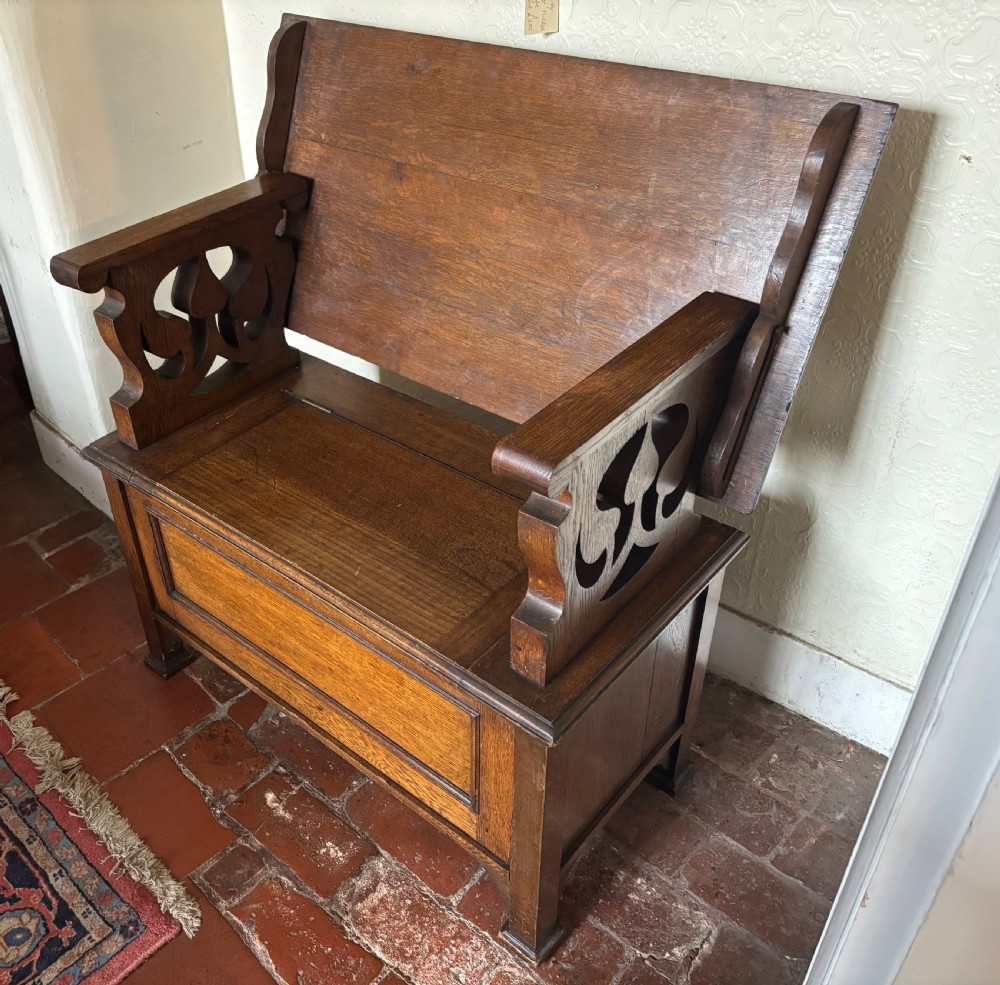 A small oak Art Nouveau monks bench with a hinged flip-top backrest, pierced heart cut-out side supports, and a hinged seat opening to a storage chest. View III