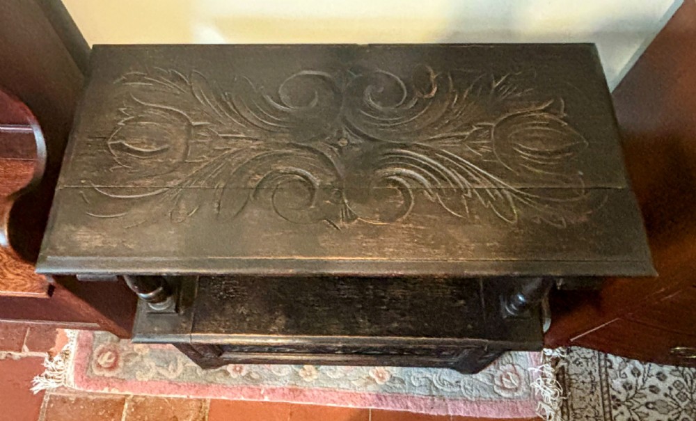 A carved dark oak monks bench featuring an upright carved backrest, turned arm supports, and a lower storage coffer base. View IV