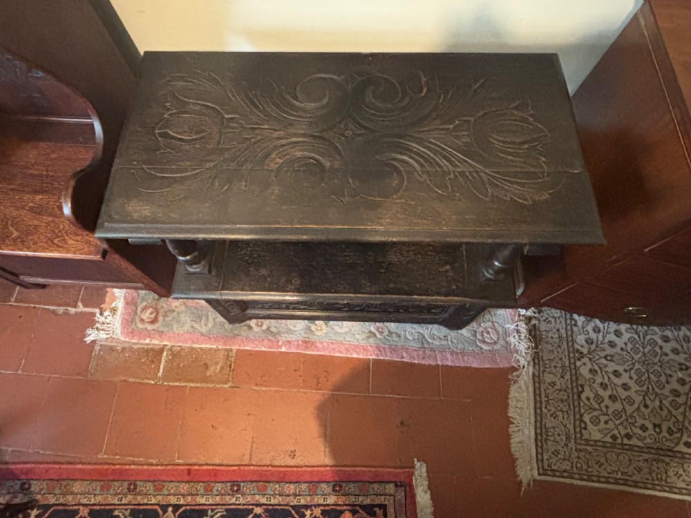A carved dark oak monks bench featuring an upright carved backrest, turned arm supports, and a lower storage coffer base. View V