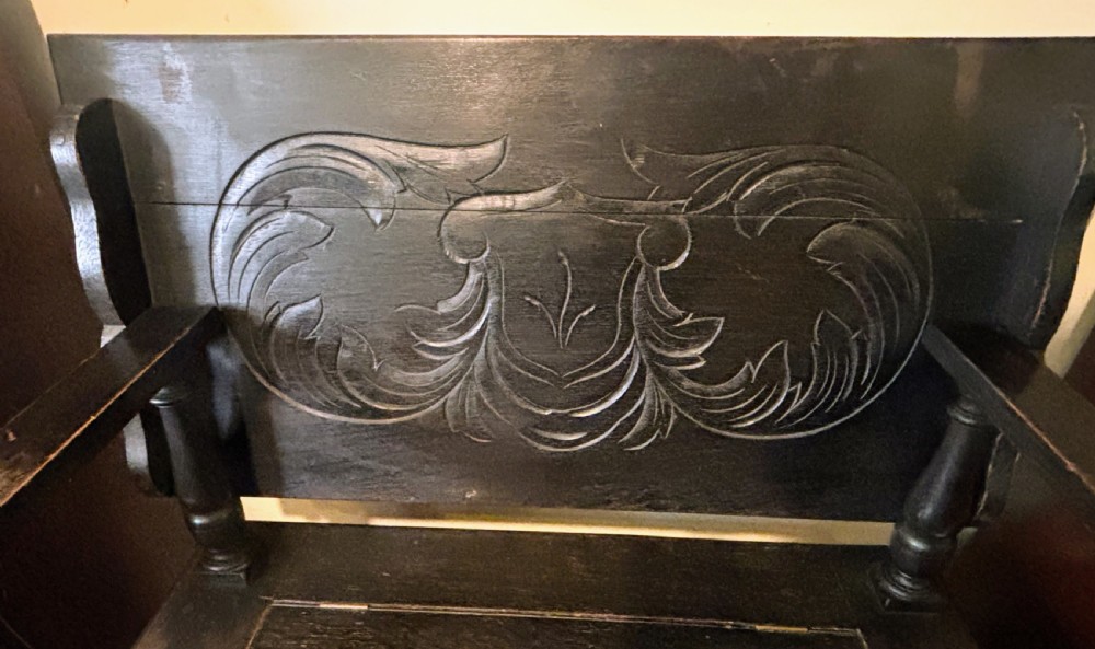A carved dark oak monks bench featuring an upright carved backrest, turned arm supports, and a lower storage coffer base. View VI