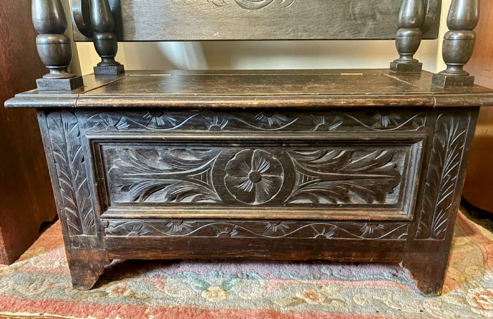 A carved dark oak monks bench featuring an upright carved backrest, turned arm supports, and a lower storage coffer base. View VII