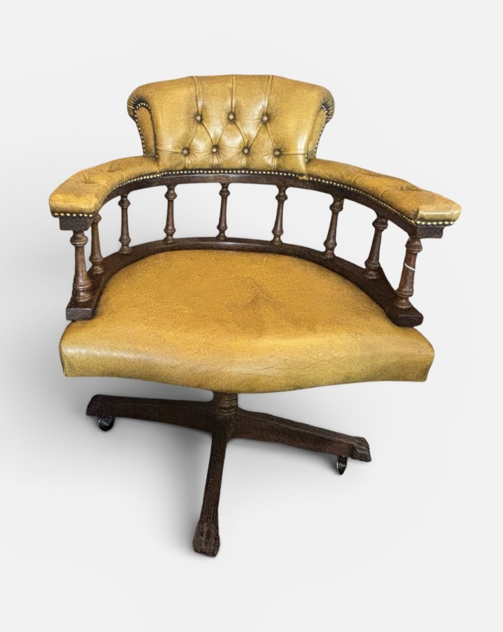 An early 20th-century mahogany revolving desk armchair featuring tan deep-buttoned leather upholstery, turned wooden spindles, brass studding, and a four-legged base on castors.