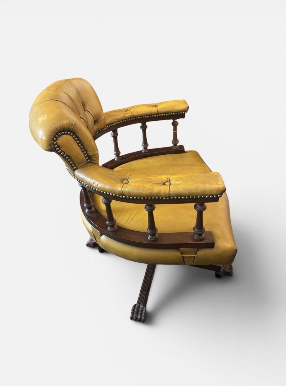 An early 20th-century mahogany revolving desk armchair featuring tan deep-buttoned leather upholstery, turned wooden spindles, brass studding, and a four-legged base on castors. View IV