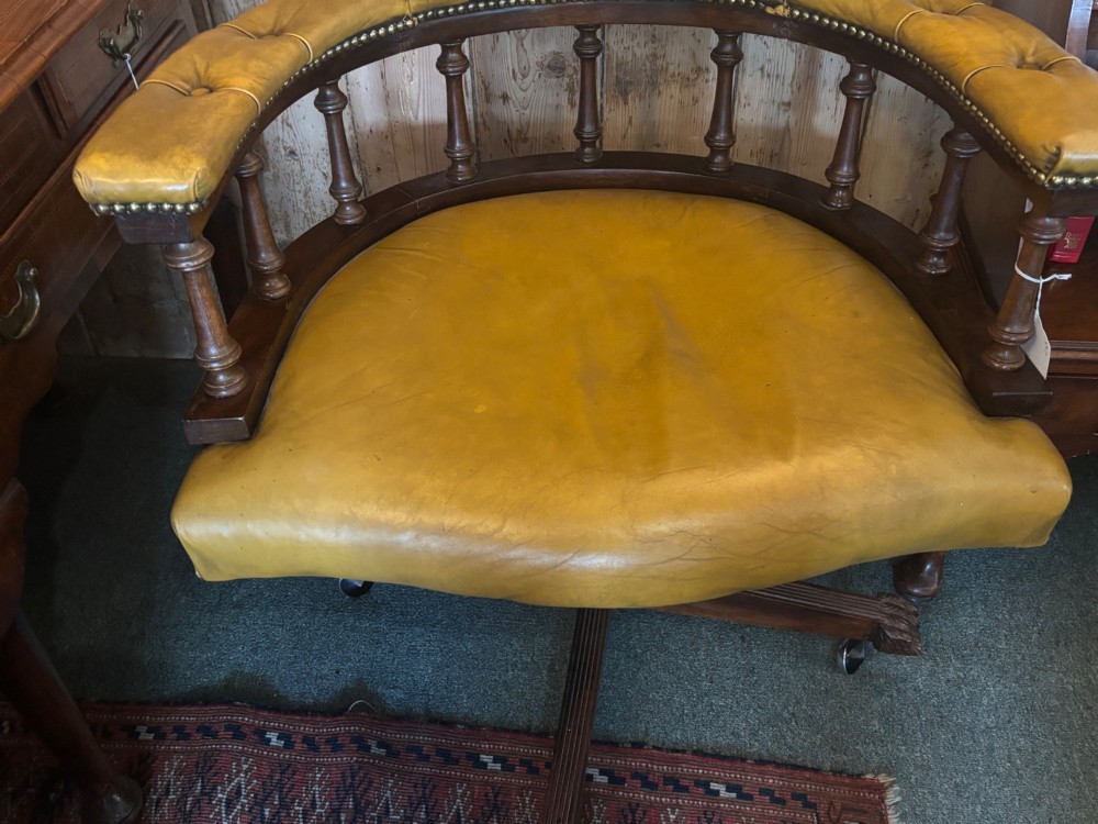 An early 20th-century mahogany revolving desk armchair featuring tan deep-buttoned leather upholstery, turned wooden spindles, brass studding, and a four-legged base on castors. View V