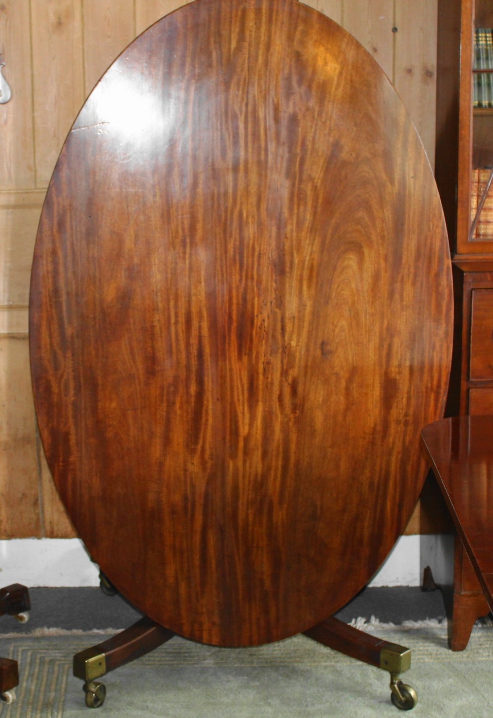 A Regency period oval mahogany breakfast table featuring a figured timber top supported by a central turned pedestal with four reeded legs ending in brass castors. View II
