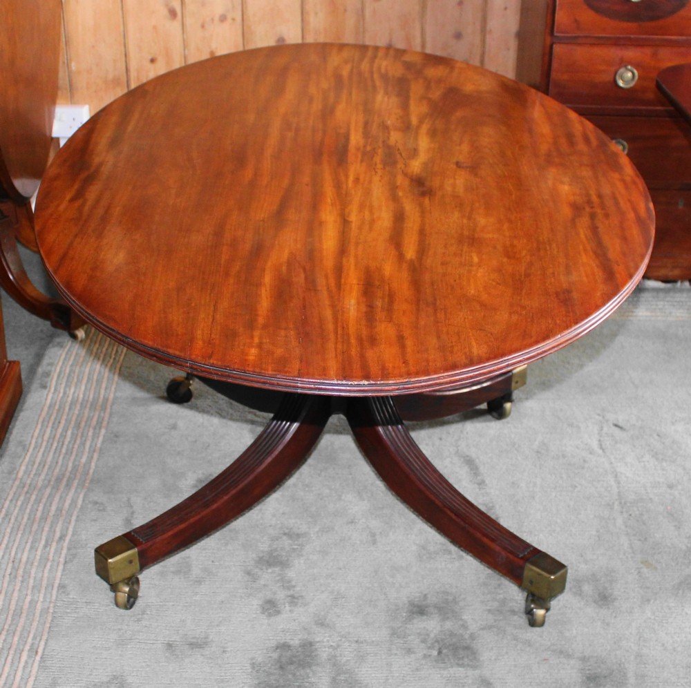 A Regency period oval mahogany breakfast table featuring a figured timber top supported by a central turned pedestal with four reeded legs ending in brass castors. View IV