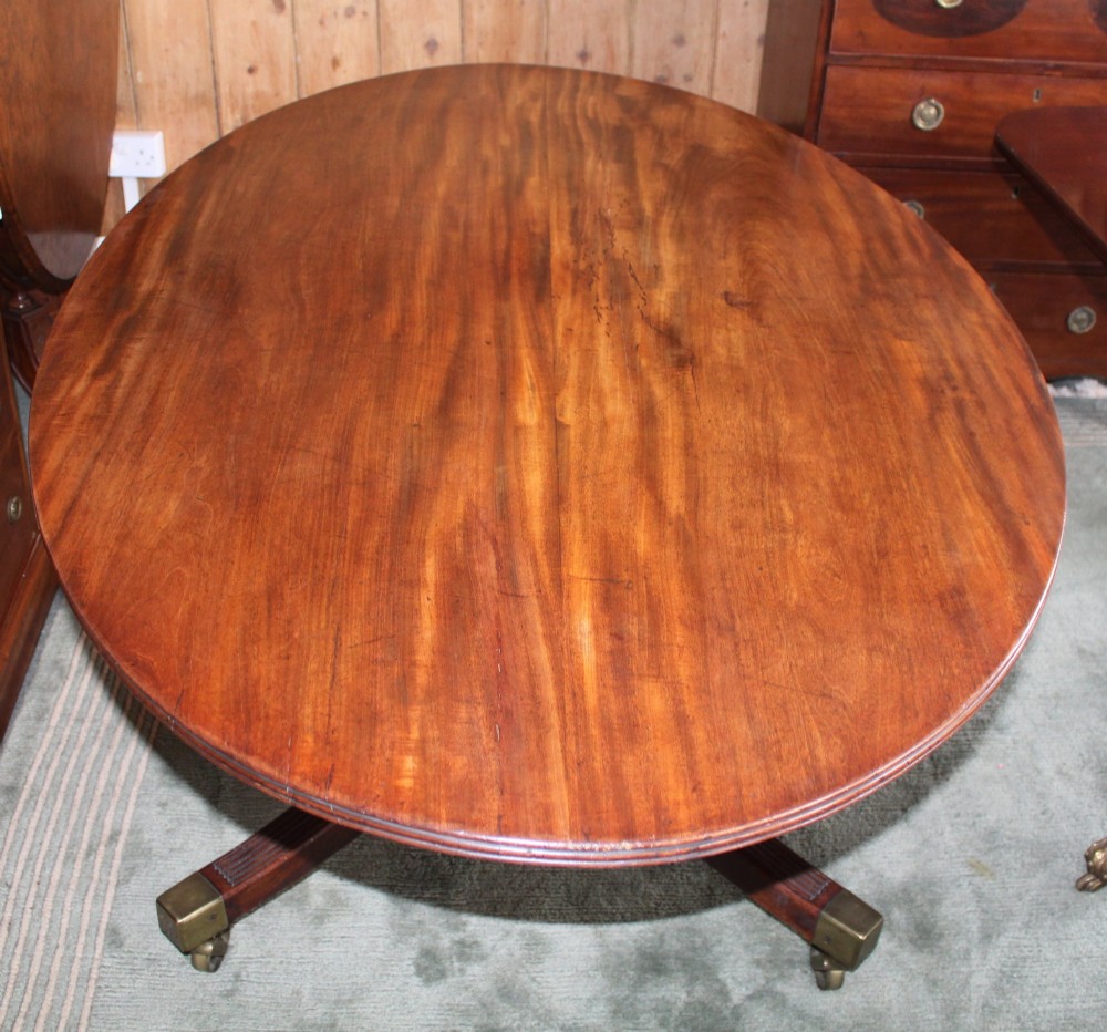 A Regency period oval mahogany breakfast table featuring a figured timber top supported by a central turned pedestal with four reeded legs ending in brass castors. View V