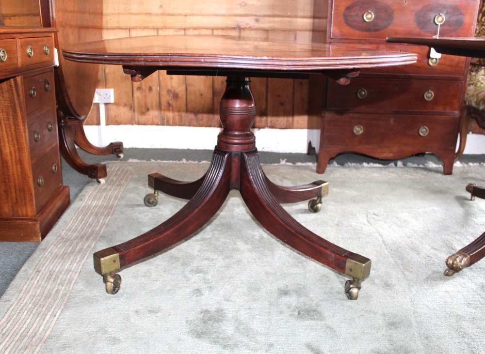 A Regency period oval mahogany breakfast table featuring a figured timber top supported by a central turned pedestal with four reeded legs ending in brass castors. View VI