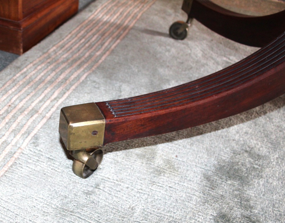 A Regency period oval mahogany breakfast table featuring a figured timber top supported by a central turned pedestal with four reeded legs ending in brass castors. View VII