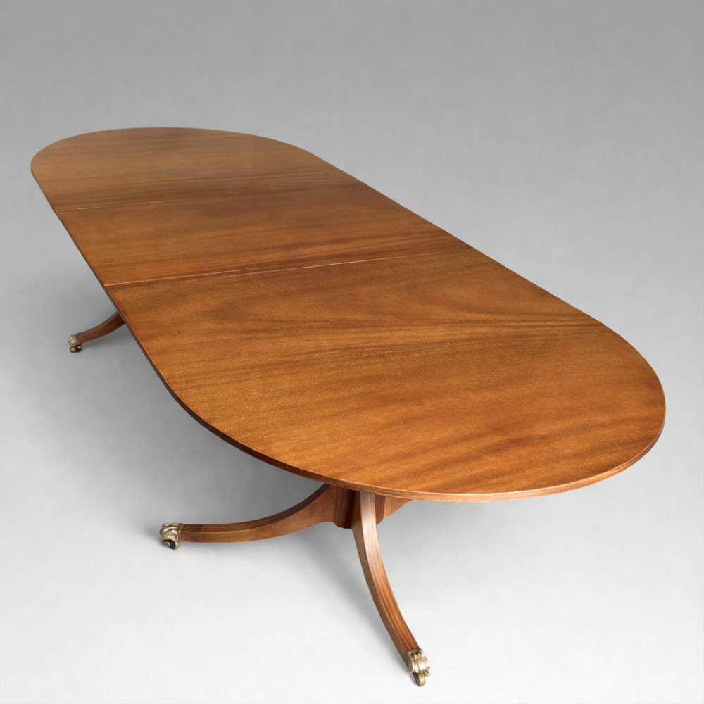 A mahogany twin pillar dining table featuring an oval top with an extension leaf, supported by two splayed pedestal bases with brass castors.