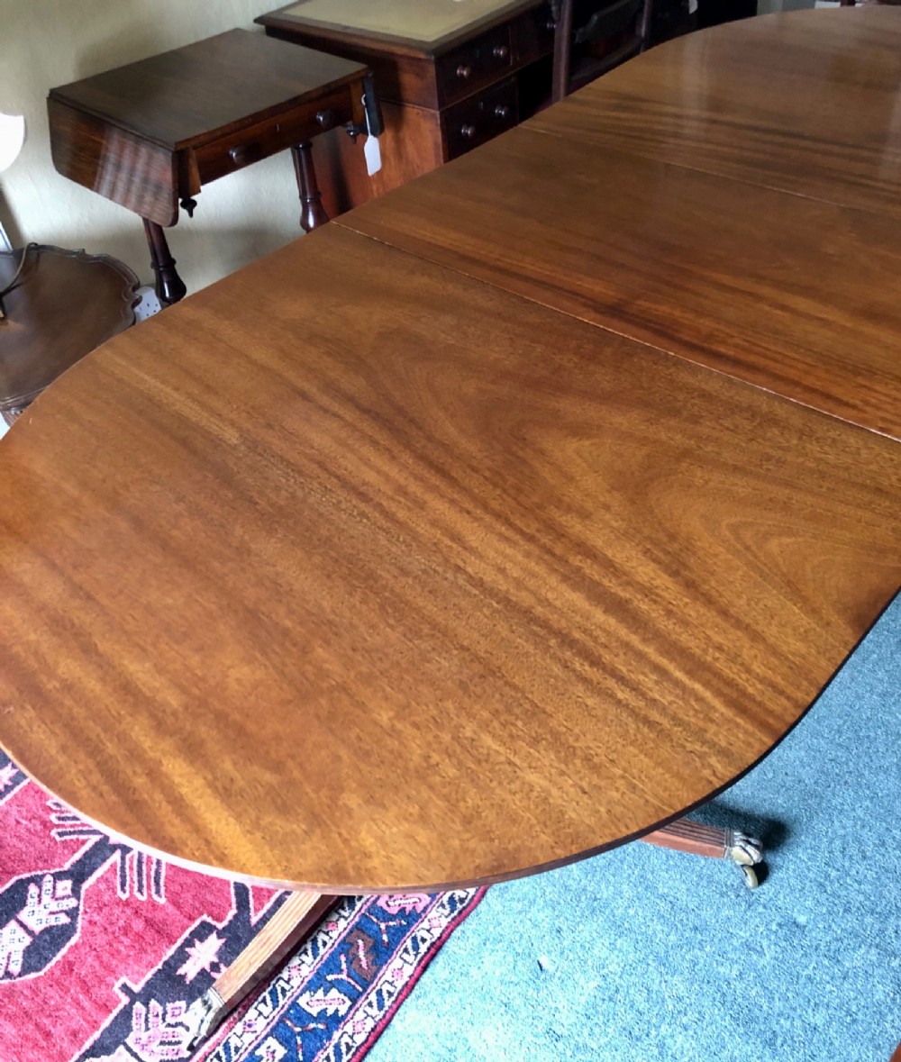 A mahogany twin pillar dining table featuring an oval top with an extension leaf, supported by two splayed pedestal bases with brass castors. View V