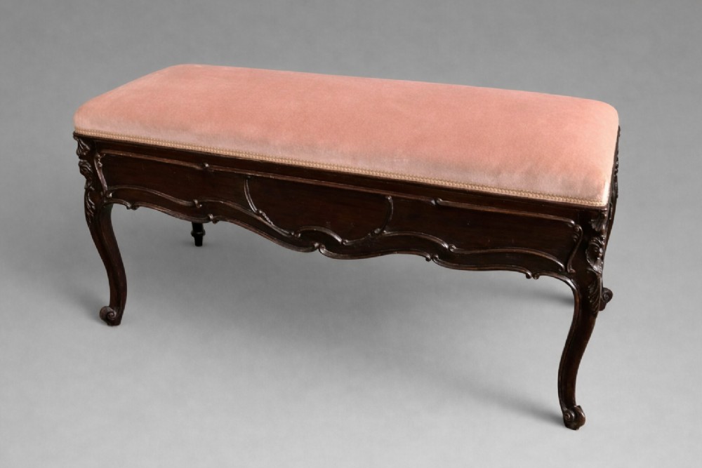An Edwardian carved rosewood duet stool with a long, dusty-pink velvet upholstered top supported by cabriole legs.