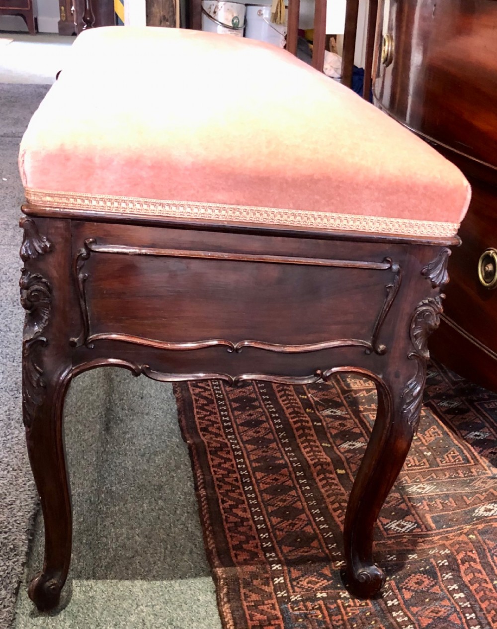 An Edwardian carved rosewood duet stool with a long, dusty-pink velvet upholstered top supported by cabriole legs. View V