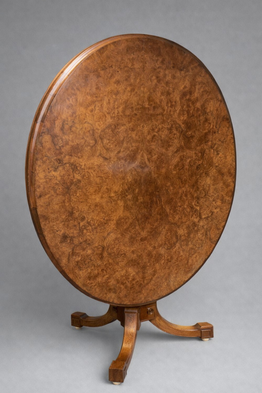 A Victorian oval burr walnut tilt-top table shown angled upright to display the wood veneer pattern and four splayed pedestal legs.