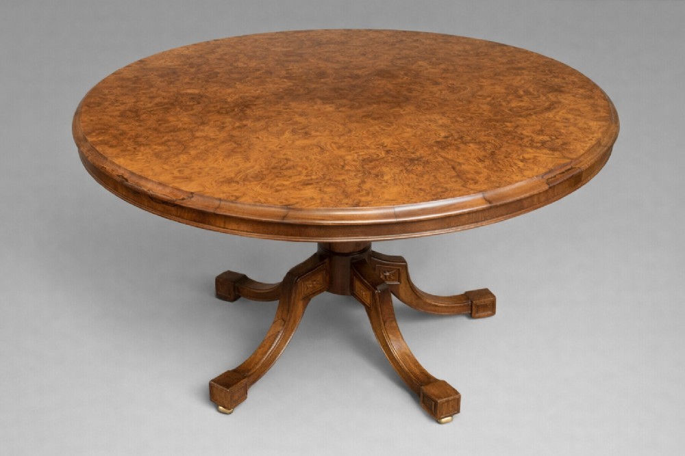 A Victorian oval burr walnut tilt-top table shown angled upright to display the wood veneer pattern and four splayed pedestal legs. View II