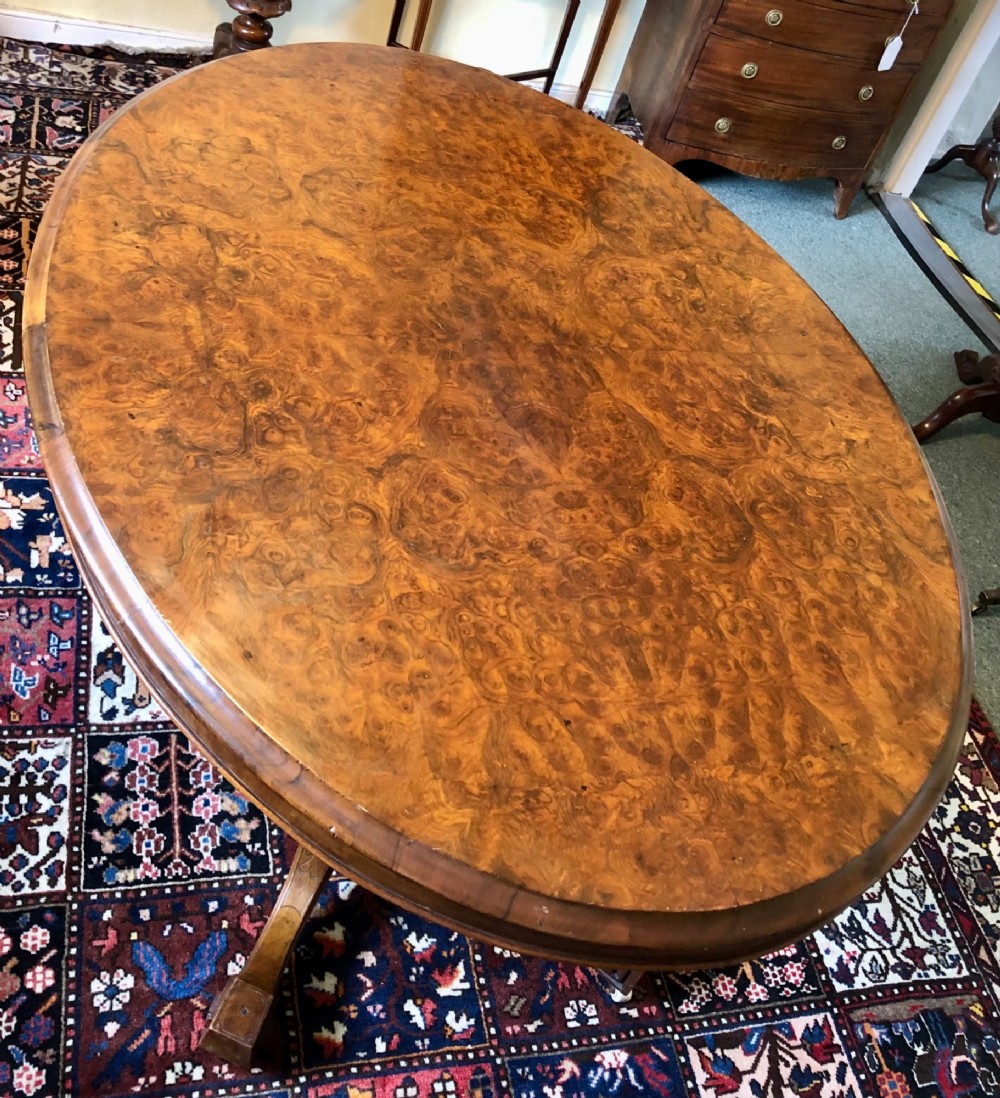 A Victorian oval burr walnut tilt-top table shown angled upright to display the wood veneer pattern and four splayed pedestal legs. View IV