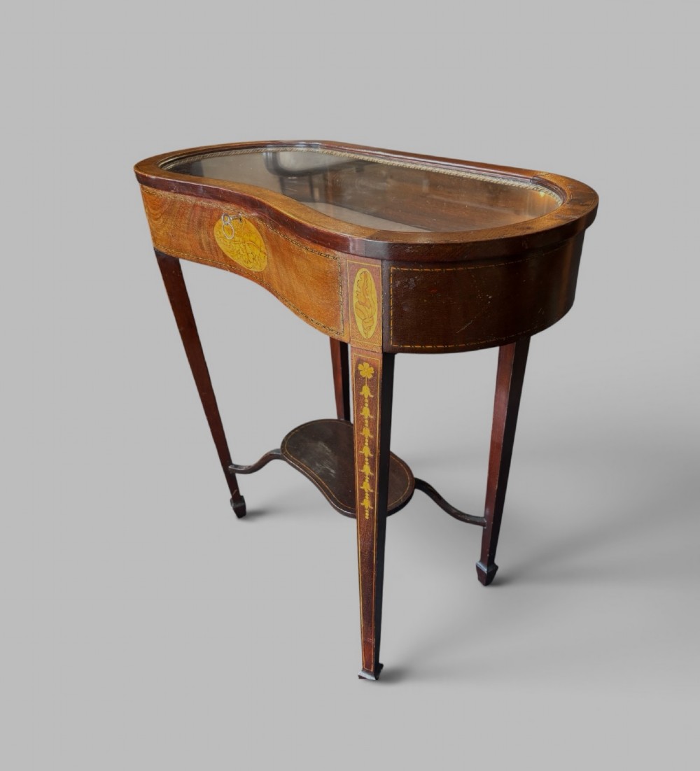 A kidney-shaped Edwardian mahogany display table with satinwood floral inlays, slender tapered legs, a glass top, and an undershelf.