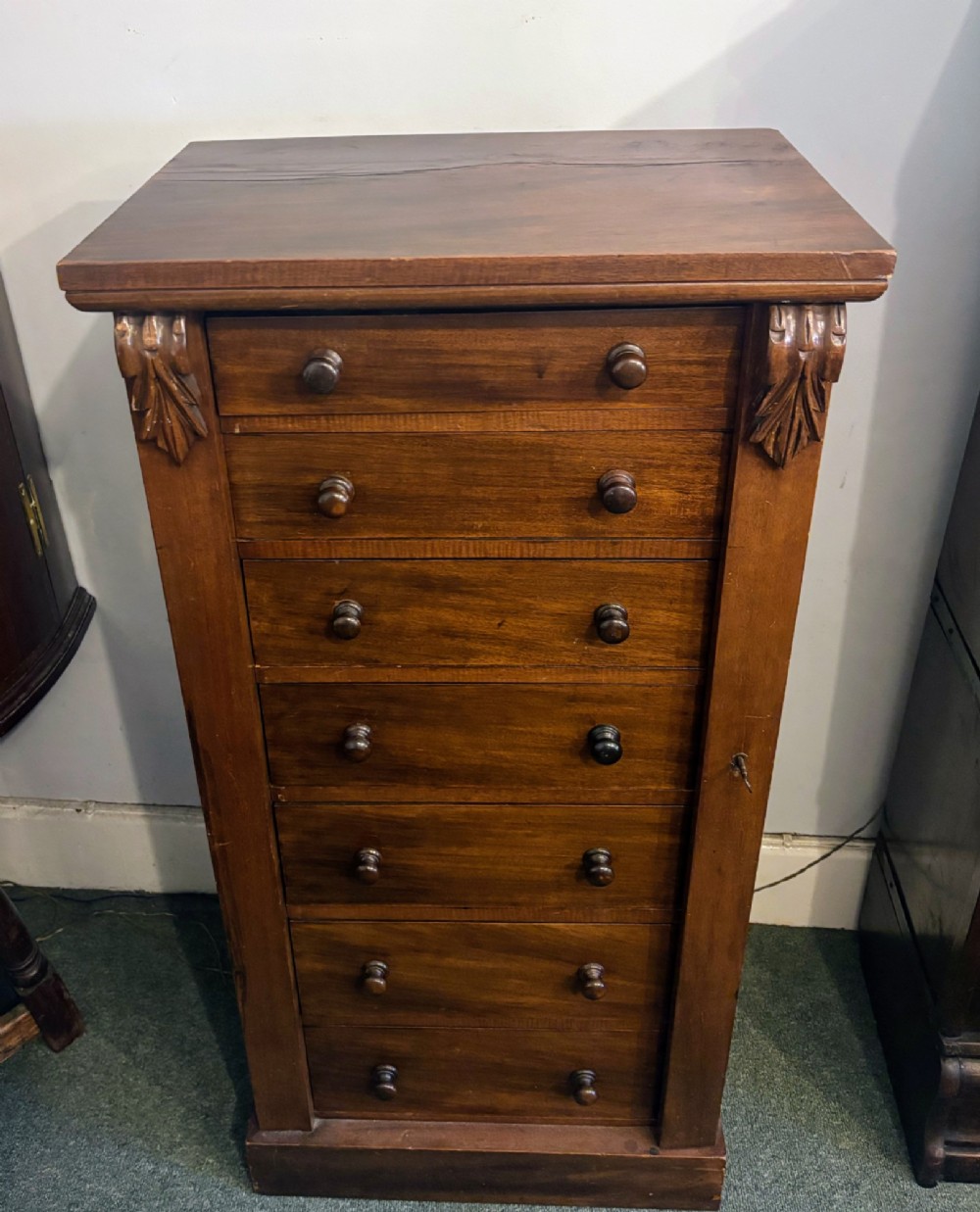 A tall Victorian walnut Wellington chest featuring seven stacked drawers, a right-hand locking stile, carved corner corbels, and turned wooden handles on a plinth base. View V
