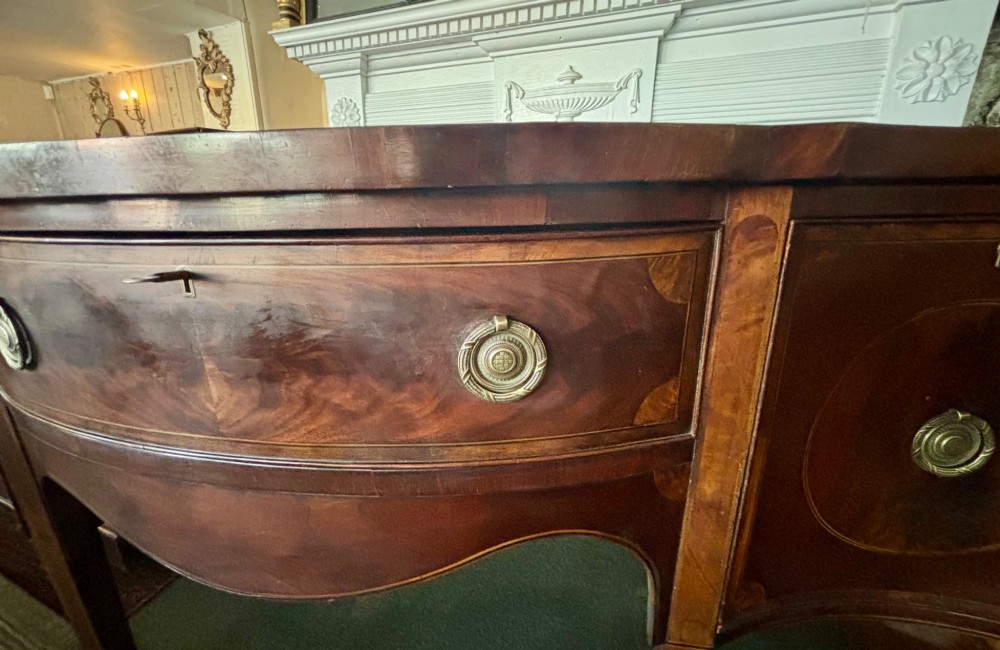 A George III mahogany serpentine-front sideboard featuring circular inlaid drawer details, brass ring handles, and square tapered legs. View IV