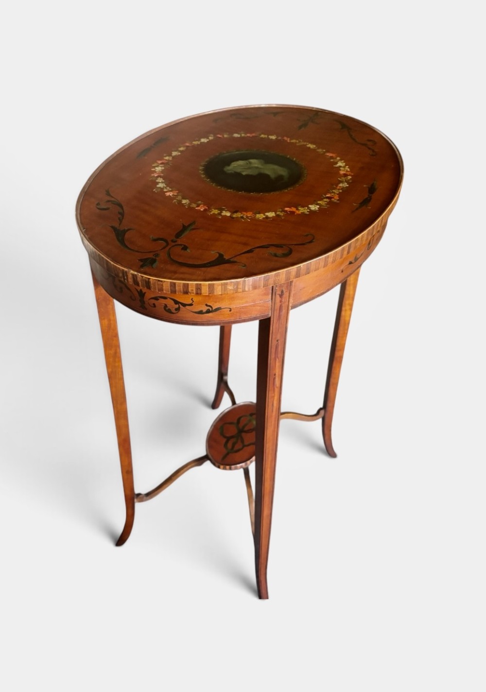An oval Edwardian satinwood occasional table featuring hand-painted floral garlands, a central classical portrait medallion, and tapered legs linked by a circular stretcher undertier. View II