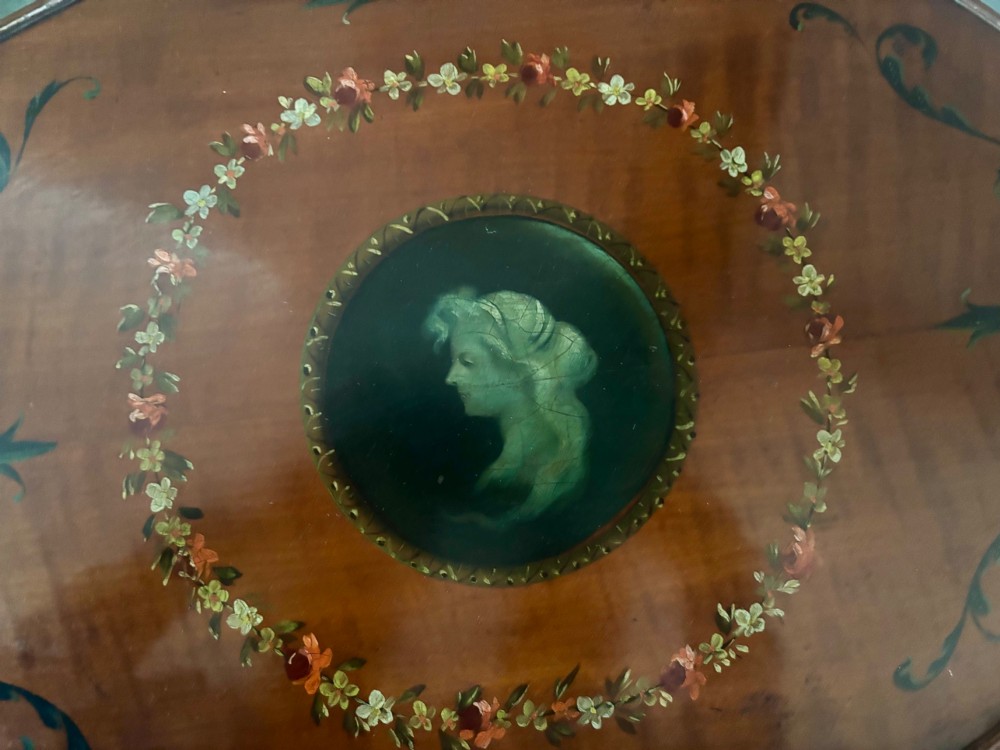 An oval Edwardian satinwood occasional table featuring hand-painted floral garlands, a central classical portrait medallion, and tapered legs linked by a circular stretcher undertier. View V