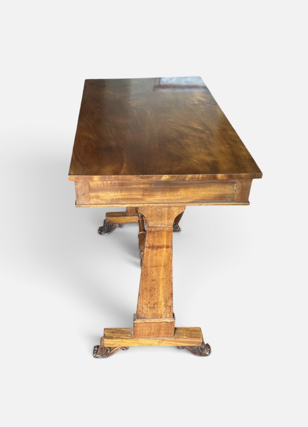 An English Regency mahogany writing table with a polished rectangular top, trestle side supports, turned central stretcher, and carved feet. View V