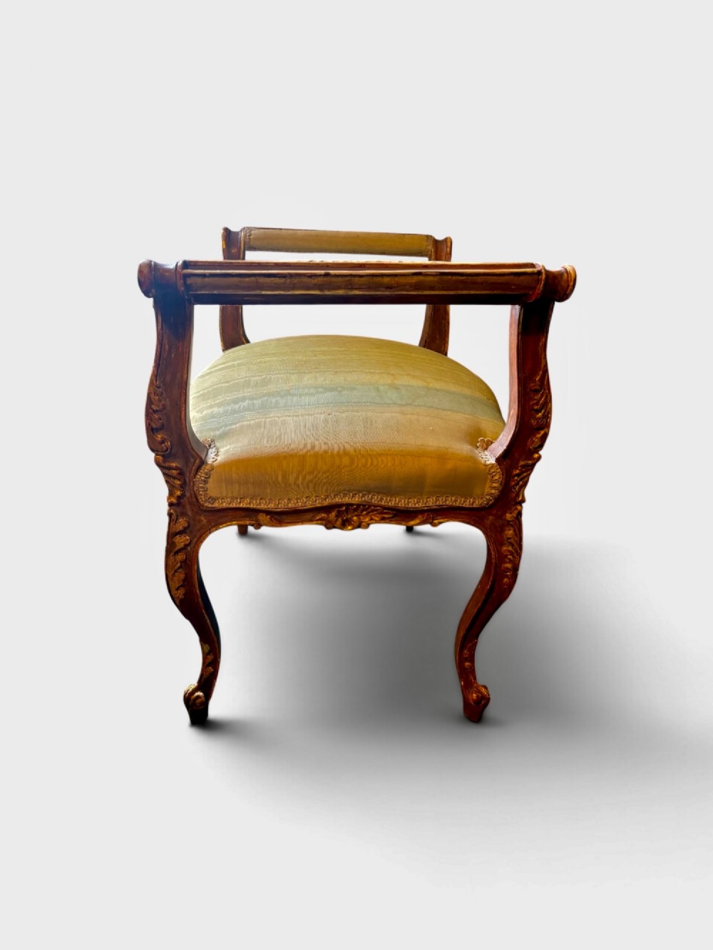 An early 20th-century Rococo Revival giltwood window seat featuring carved cabriole legs, padded raised scroll arms, and olive green striped silk upholstery. View II