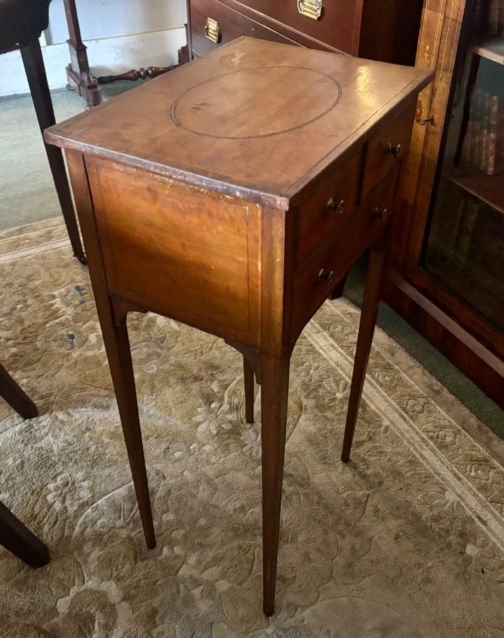 An early nineteenth-century satinwood side table featuring an oval inlaid top panel, three drawers with brass knob handles, and slender tapered square legs. View V