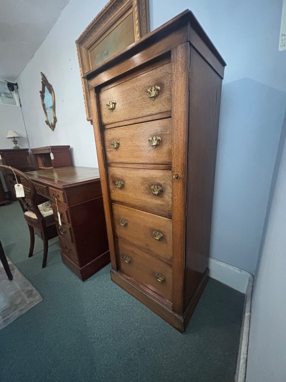 A tall, narrow Edwardian mahogany Wellington chest featuring five graduated drawers with brass handles and a hinged side locking bar on a plinth base. View VI