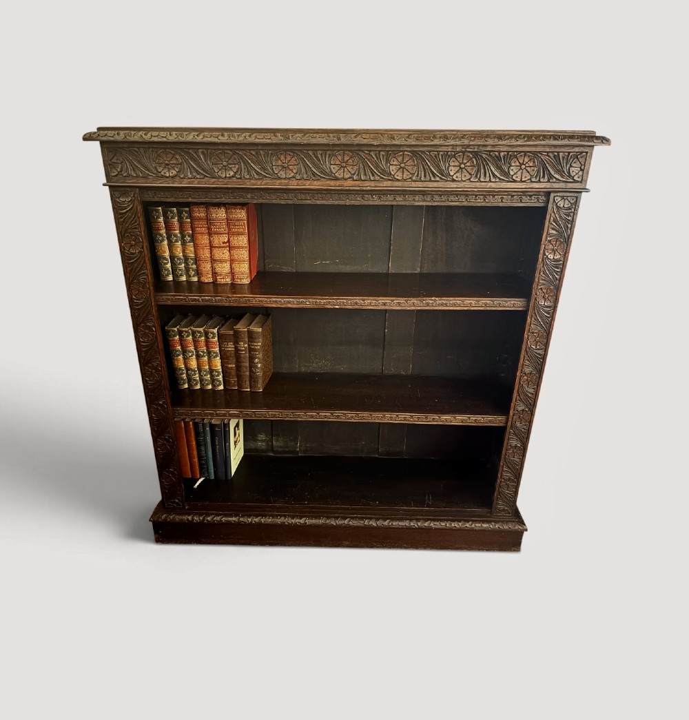 A Victorian dark carved oak open bookcase featuring detailed rosette hand-carvings along the frieze and outer border with three open shelves holding books.