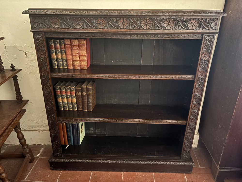 A Victorian dark carved oak open bookcase featuring detailed rosette hand-carvings along the frieze and outer border with three open shelves holding books. View V