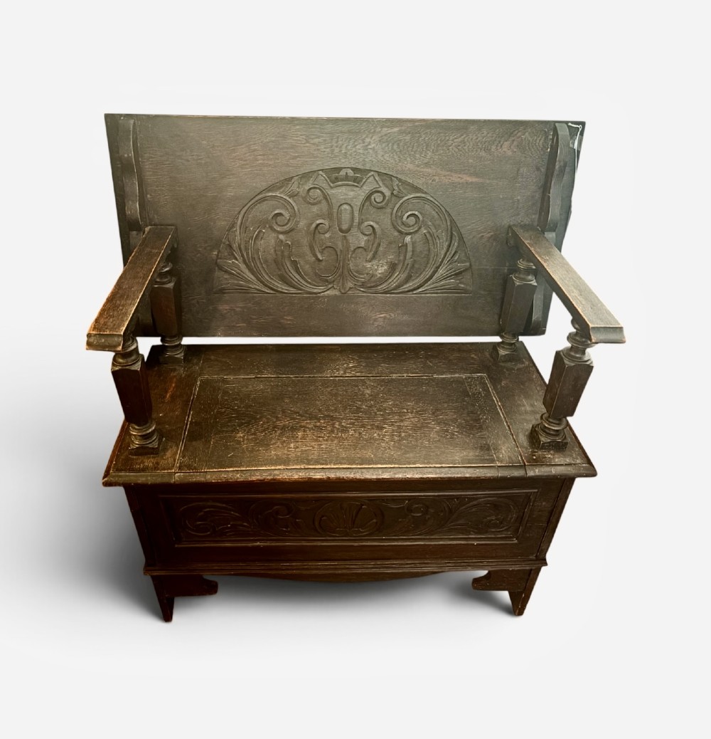 A dark carved oak monks bench featuring a pivoted panel backrest, turned arm supports, and a chest storage base.