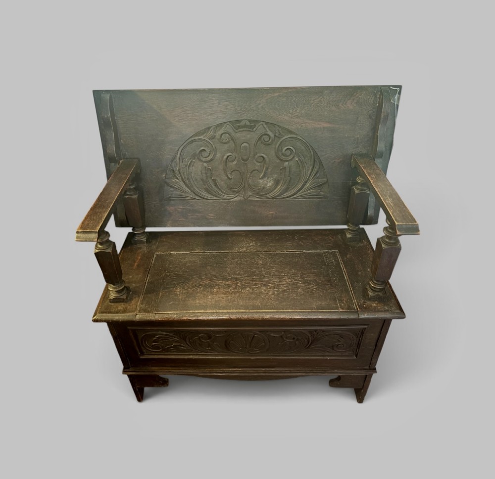 A dark carved oak monks bench featuring a pivoted panel backrest, turned arm supports, and a chest storage base. View II