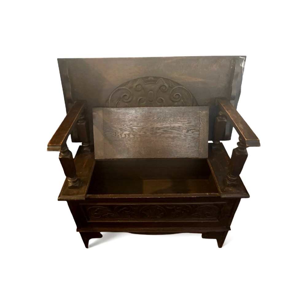 A dark carved oak monks bench featuring a pivoted panel backrest, turned arm supports, and a chest storage base. View III