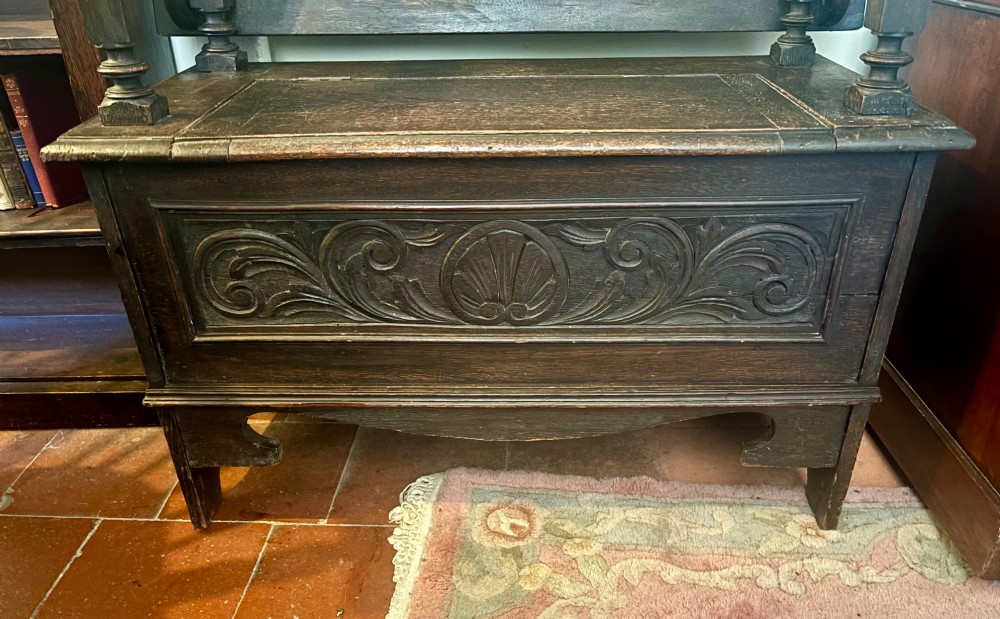 A dark carved oak monks bench featuring a pivoted panel backrest, turned arm supports, and a chest storage base. View V