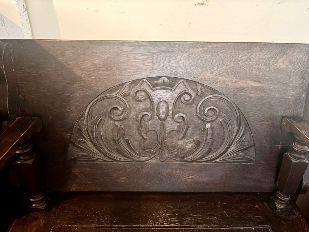 A dark carved oak monks bench featuring a pivoted panel backrest, turned arm supports, and a chest storage base. View VI