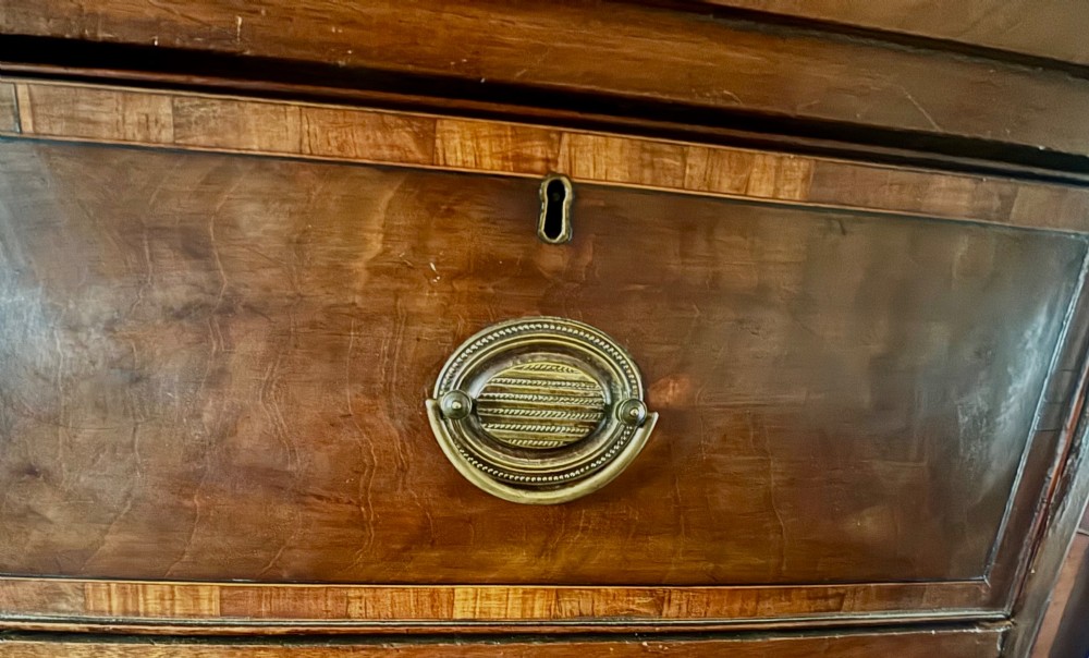 A Regency mahogany bowfront chest of drawers featuring satinwood crossbanding, brass oval handles, and splayed legs. View IV