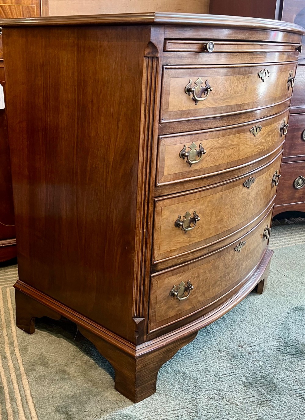 A burr walnut bow-front chest of four graduated drawers featuring brass drop handles, herringbone crossbanding, and bracket feet. View VIII